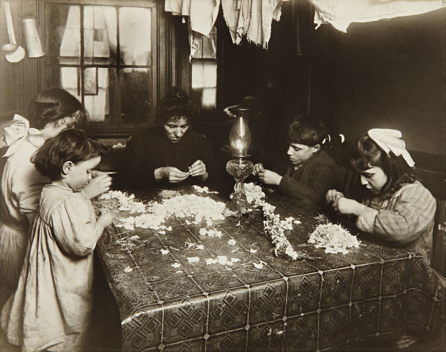 Lewis Hine — This was the high-tide of tenement homework in New York City when children like these worked at flower-making, picking out nut-meats and many other and many other things long days and even into the night,(this was going on at 8;00 P.M.) Their schooling, health and recreation suffered very much.